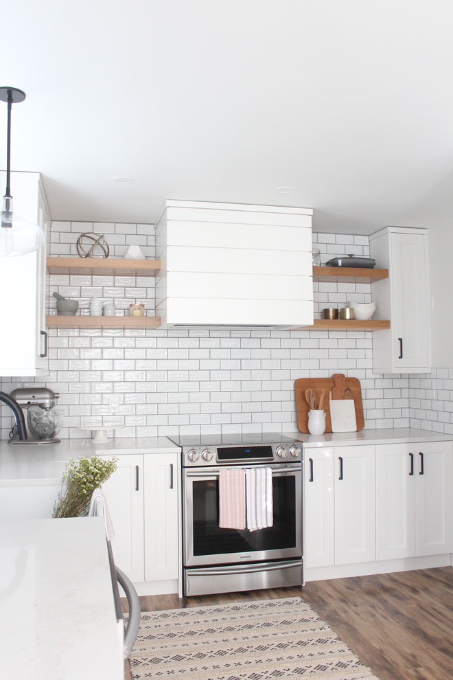 This is an example of a mid-sized country l-shaped separate kitchen in Vancouver with a farmhouse sink, shaker cabinets, white cabinets, solid surface benchtops, white splashback, ceramic splashback, stainless steel appliances, laminate floors, no island, brown floor and white benchtop.