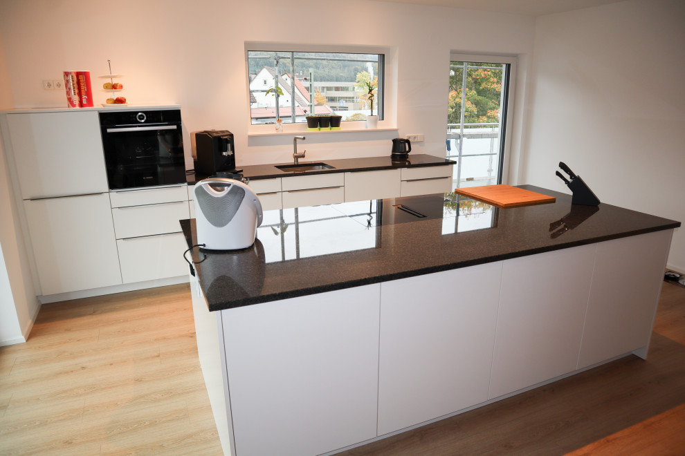 Küche in L-Form mit Einbauwaschbecken, Kassettenfronten, weißen Schränken, Mineralwerkstoff-Arbeitsplatte, Küchenrückwand in Weiß, Küchengeräten aus Edelstahl, Laminat, Kücheninsel, braunem Boden, schwarzer Arbeitsplatte und kleiner Kücheninsel