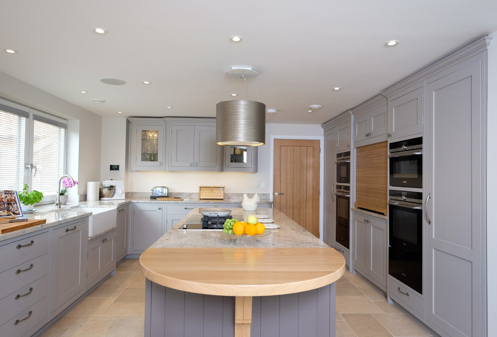Design ideas for a large transitional u-shaped open plan kitchen in Hampshire with a farmhouse sink, shaker cabinets, grey cabinets, granite benchtops, black appliances, limestone floors and with island.