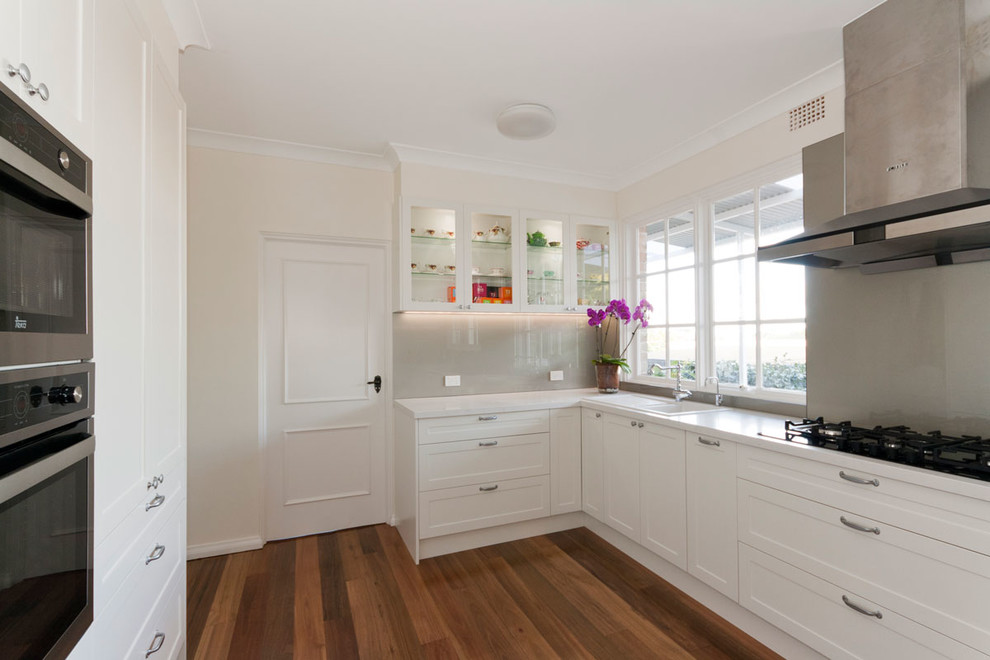 This is an example of a mid-sized traditional u-shaped kitchen in Sydney with a single-bowl sink, shaker cabinets, white cabinets, quartz benchtops, glass sheet splashback, medium hardwood floors and no island.