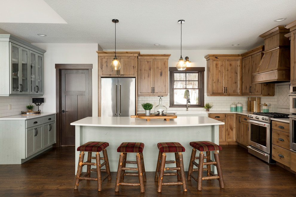 Country l-shaped kitchen in Minneapolis with a farmhouse sink, shaker cabinets, medium wood cabinets, white splashback, subway tile splashback, stainless steel appliances, dark hardwood floors, with island, brown floor and white benchtop.
