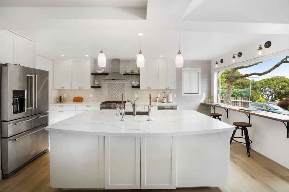 Mid-sized beach style open plan kitchen in San Francisco with an undermount sink, shaker cabinets, white cabinets, quartz benchtops, grey splashback, ceramic splashback, stainless steel appliances, light hardwood floors, with island and white benchtop.