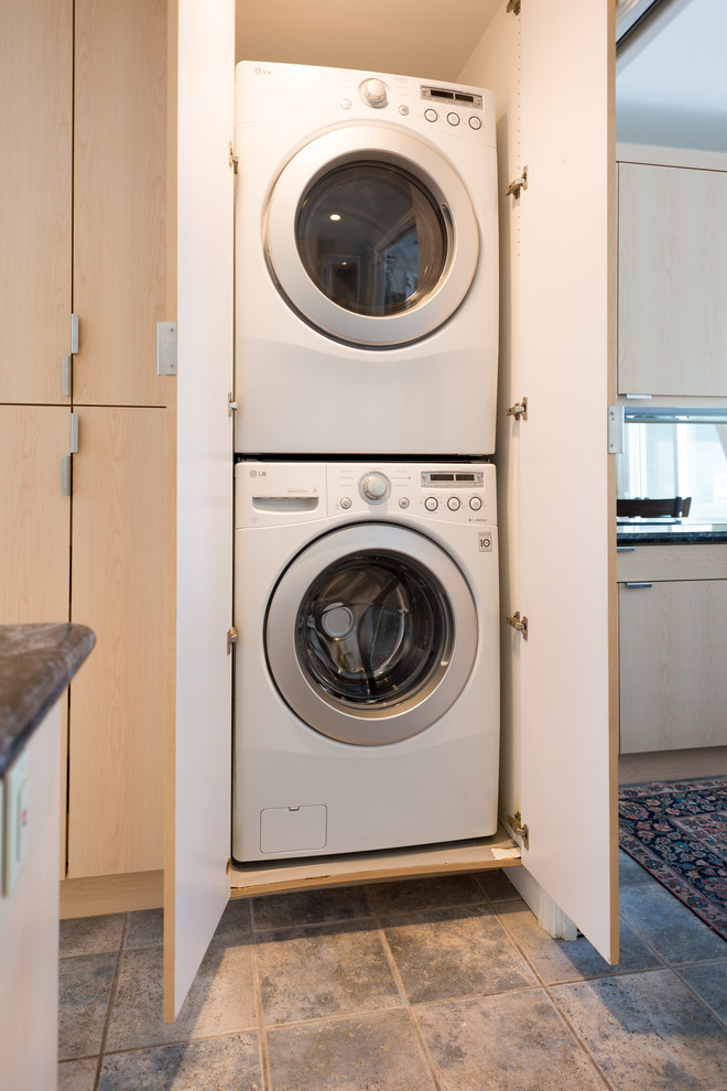 Photo of a small beach style laundry cupboard in San Diego with flat-panel cabinets, light wood cabinets, travertine floors and a stacked washer and dryer.