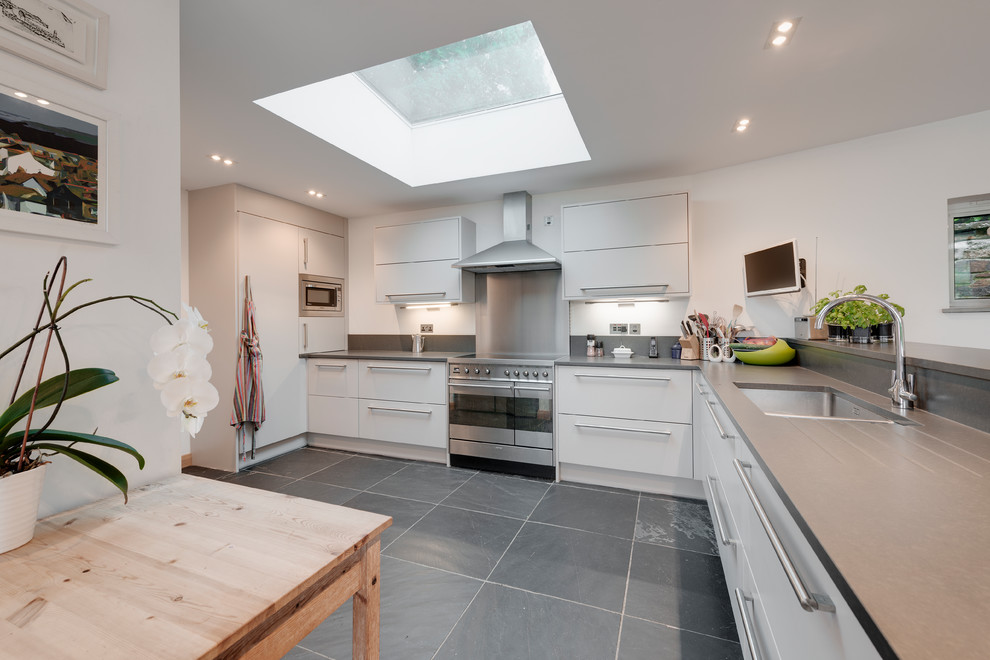This is an example of a mid-sized modern u-shaped open plan kitchen in Cornwall with an integrated sink, flat-panel cabinets, white cabinets, solid surface benchtops, grey splashback, slate splashback, stainless steel appliances, slate floors, with island and grey floor.