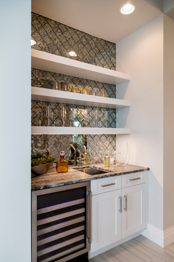 This is an example of a small transitional single-wall wet bar in Miami with an undermount sink, shaker cabinets, white cabinets, granite benchtops, multi-coloured splashback, mirror splashback, ceramic floors, beige floor and grey benchtop.