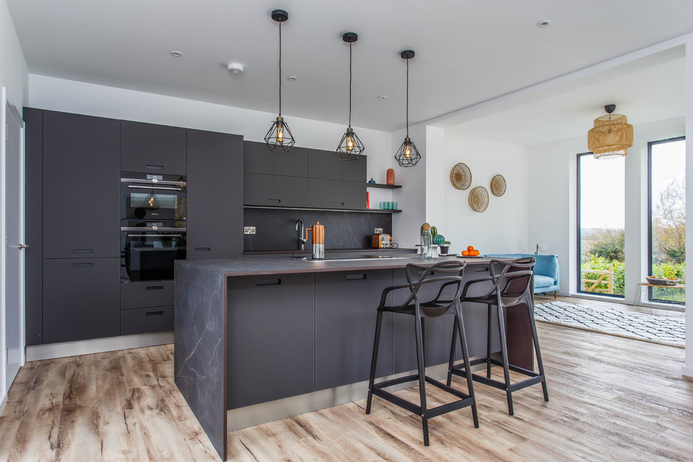 Design ideas for a contemporary galley kitchen in Sussex with flat-panel cabinets, black cabinets, black splashback, black appliances, medium hardwood floors, with island, brown floor and black benchtop.