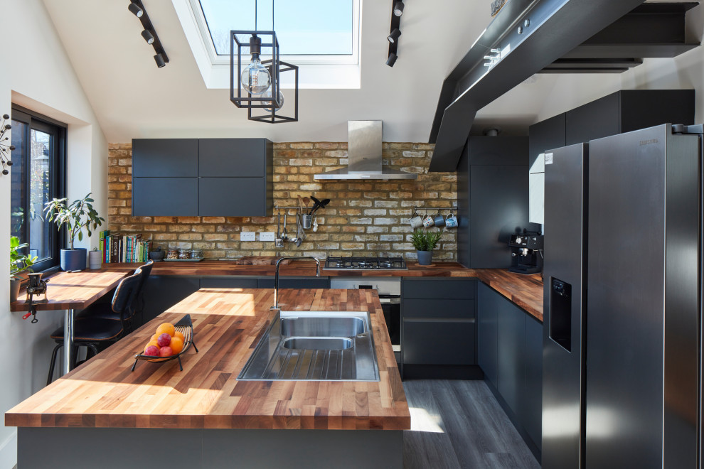 Photo of a large contemporary u-shaped open plan kitchen in London with a drop-in sink, flat-panel cabinets, wood benchtops, stainless steel appliances, light hardwood floors, with island, brown floor, brown benchtop, black cabinets, brown splashback and brick splashback.