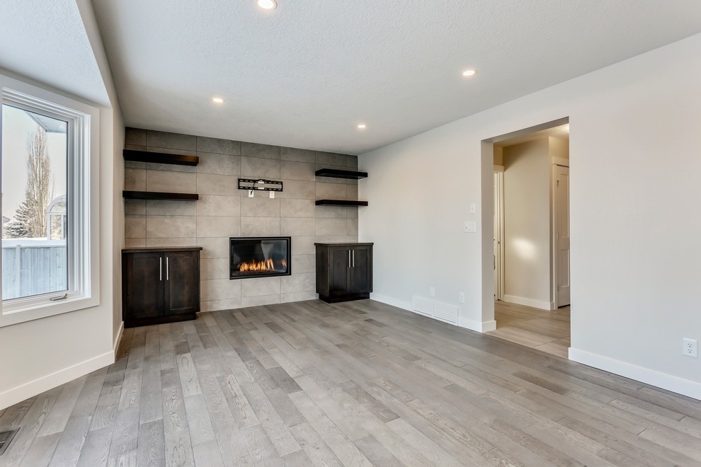 Photo of a large transitional open concept living room in Calgary with white walls, medium hardwood floors, a standard fireplace, a tile fireplace surround, a wall-mounted tv and brown floor.