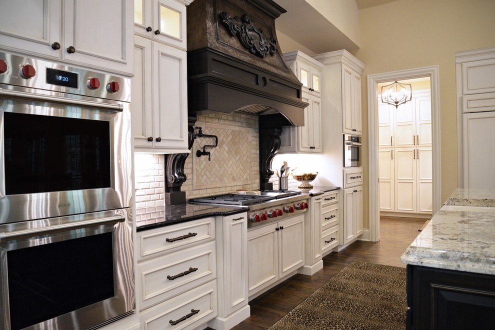 Photo of a large traditional l-shaped kitchen in St Louis with a farmhouse sink, recessed-panel cabinets, white cabinets, granite benchtops, beige splashback, stone tile splashback, stainless steel appliances, medium hardwood floors, with island, brown floor and black benchtop.