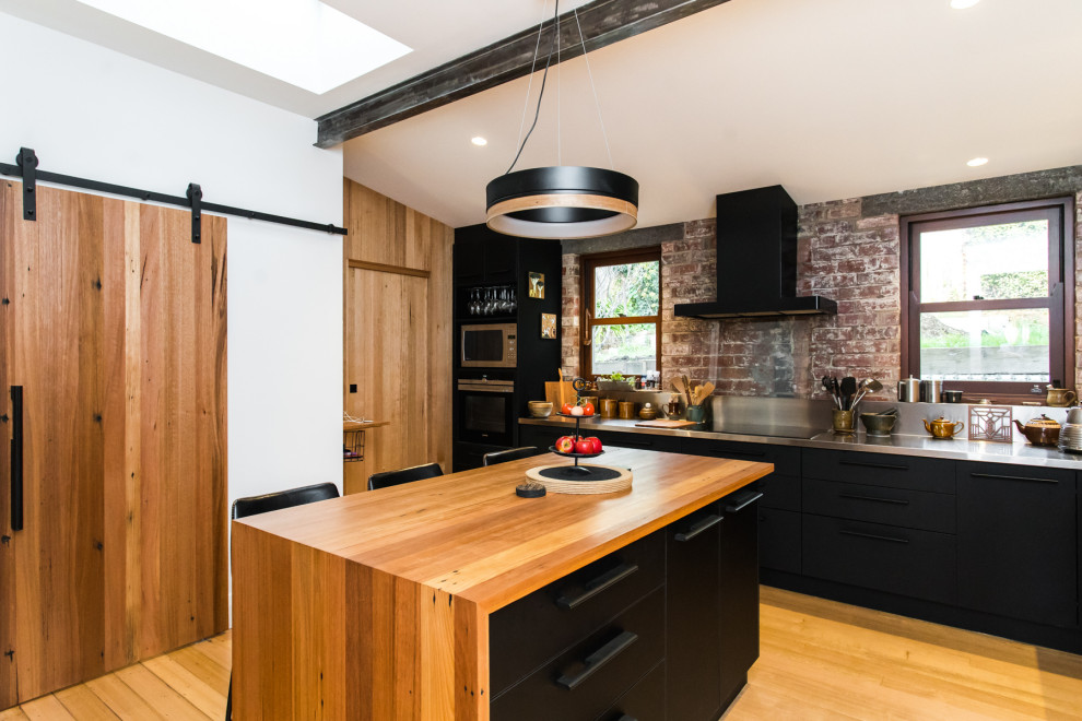 Inspiration for an industrial kitchen in Hobart with flat-panel cabinets, black cabinets, stainless steel benchtops, brick splashback, light hardwood floors, with island and beige floor.