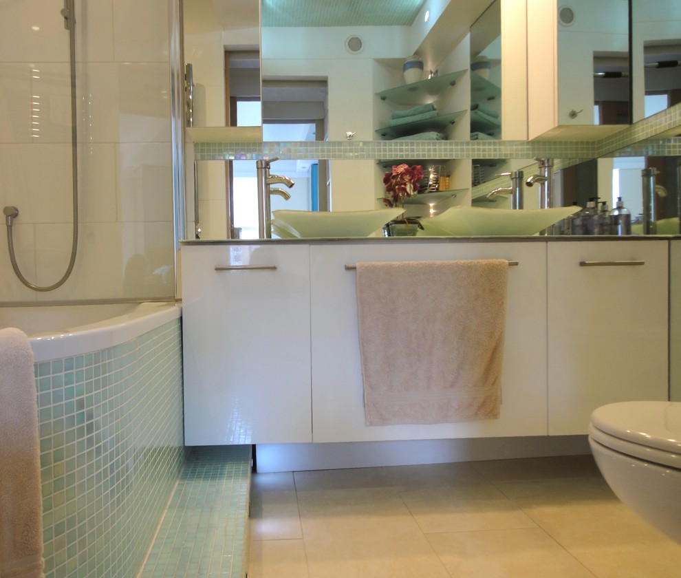 Photo of a small modern bathroom in London with a vessel sink, flat-panel cabinets, white cabinets, glass benchtops, an alcove tub, a shower/bathtub combo, a wall-mount toilet, green tile, mosaic tile, white walls and ceramic floors.