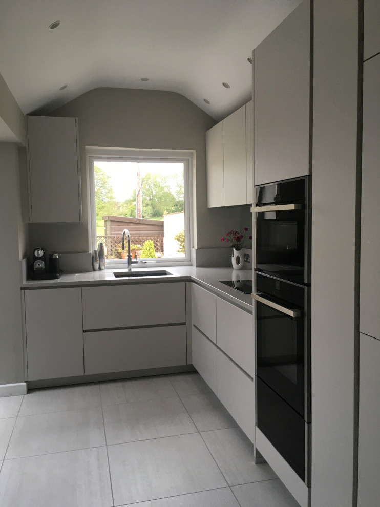 Design ideas for a small contemporary l-shaped eat-in kitchen in Manchester with an undermount sink, flat-panel cabinets, white cabinets, quartzite benchtops, metallic splashback, mirror splashback, black appliances, porcelain floors, no island, grey floor, white benchtop and vaulted.