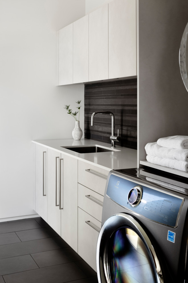 Modern laundry room in DC Metro with an undermount sink, flat-panel cabinets, grey cabinets, black walls, a side-by-side washer and dryer, black floor and white benchtop.
