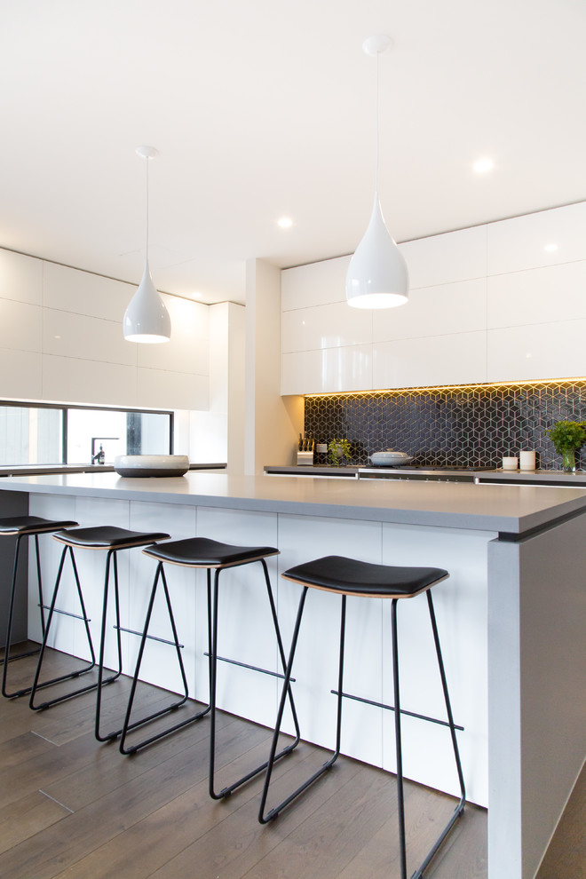 Photo of a mid-sized contemporary galley open plan kitchen in Melbourne with a drop-in sink, flat-panel cabinets, white cabinets, concrete benchtops, black splashback, ceramic splashback, stainless steel appliances, dark hardwood floors, with island and grey benchtop.