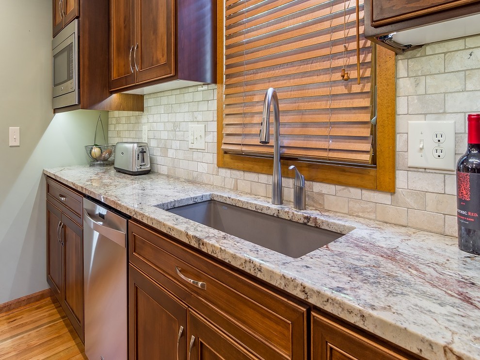 Photo of a mid-sized traditional l-shaped separate kitchen in Kansas City with an undermount sink, flat-panel cabinets, brown cabinets, granite benchtops, beige splashback, stone tile splashback, stainless steel appliances, light hardwood floors and with island.