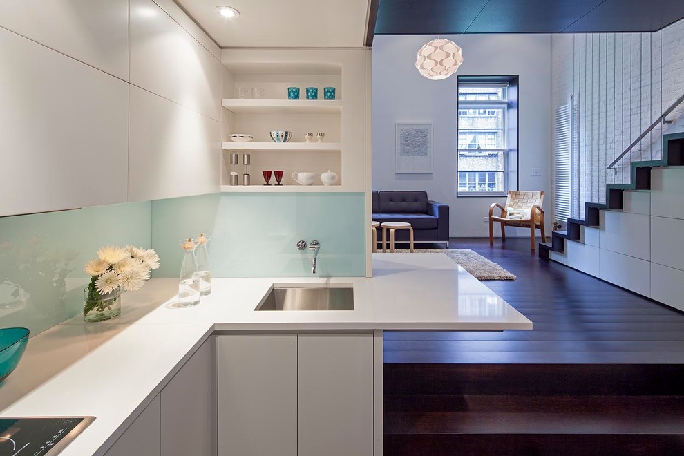 This is an example of a small modern l-shaped open plan kitchen in New York with an undermount sink, flat-panel cabinets, white cabinets, glass sheet splashback, dark hardwood floors, stainless steel appliances, white splashback and a peninsula.