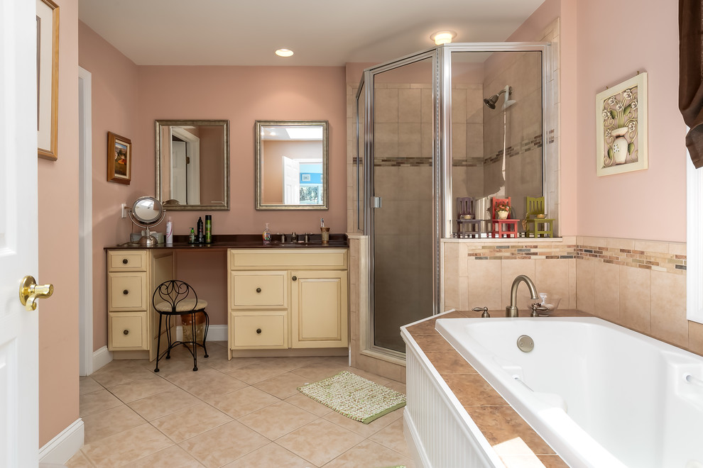 This is an example of a mid-sized beach style master bathroom in Philadelphia with raised-panel cabinets, beige cabinets, a drop-in tub, a corner shower, beige tile, ceramic tile, pink walls, ceramic floors, an undermount sink, beige floor and a hinged shower door.