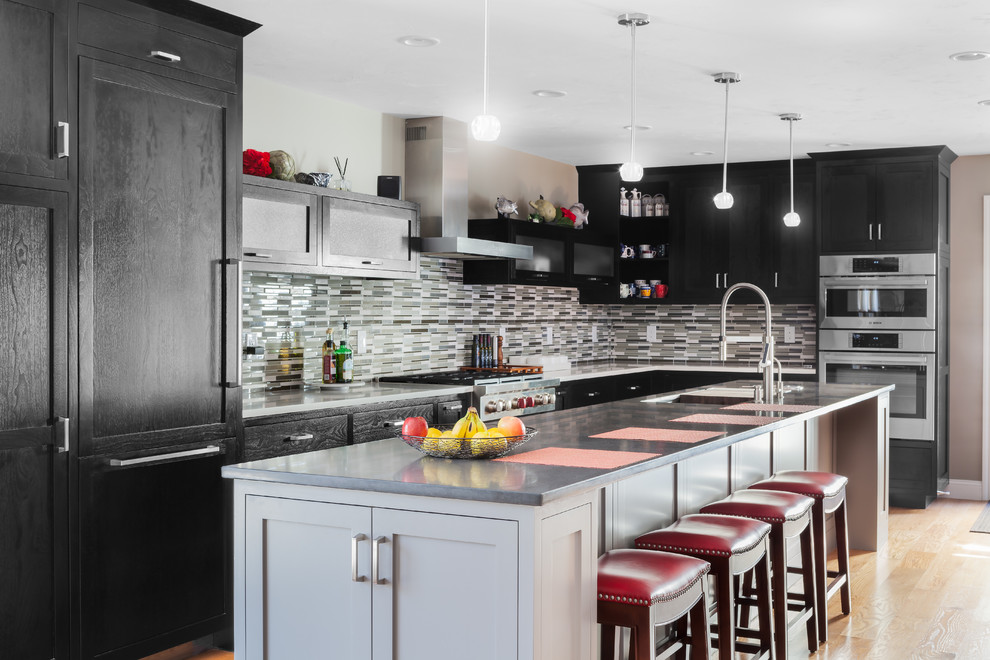 This is an example of a large transitional l-shaped eat-in kitchen in Boston with shaker cabinets, black cabinets, matchstick tile splashback, stainless steel appliances, with island, a double-bowl sink, solid surface benchtops, multi-coloured splashback, light hardwood floors and brown floor.