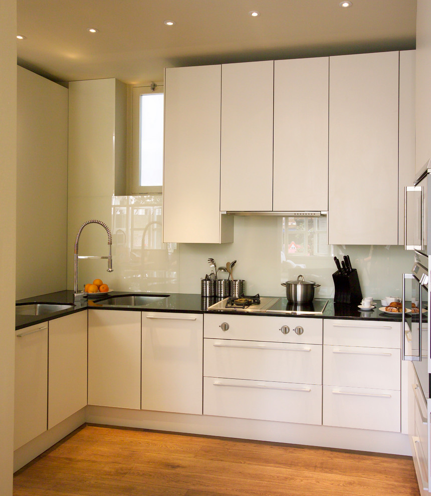 This is an example of a small contemporary u-shaped separate kitchen in Berkshire with an undermount sink, flat-panel cabinets, white cabinets, white splashback, glass sheet splashback, medium hardwood floors and no island.