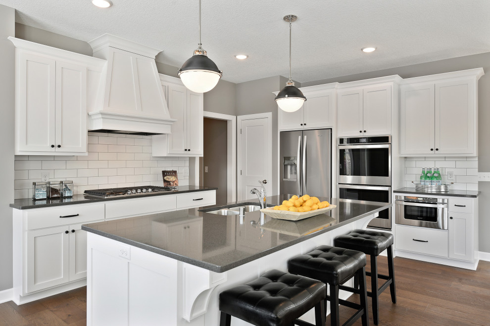 Example of a large transitional l-shaped medium tone wood floor and brown floor kitchen design in Minneapolis with a double-bowl sink, shaker cabinets, white cabinets, white backsplash, subway tile backsplash, stainless steel appliances, an island and gray countertops