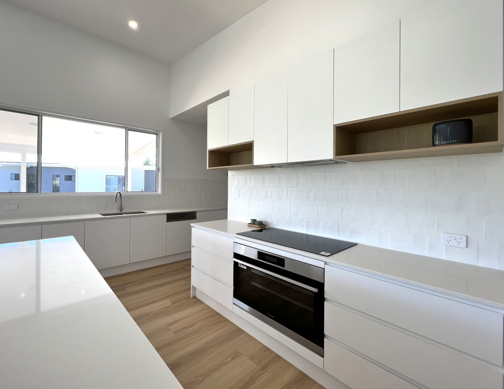 Inspiration for a contemporary l-shaped open plan kitchen in Sunshine Coast with an undermount sink, flat-panel cabinets, white cabinets, quartz benchtops, white splashback, ceramic splashback, stainless steel appliances, vinyl floors, with island, white benchtop and vaulted.