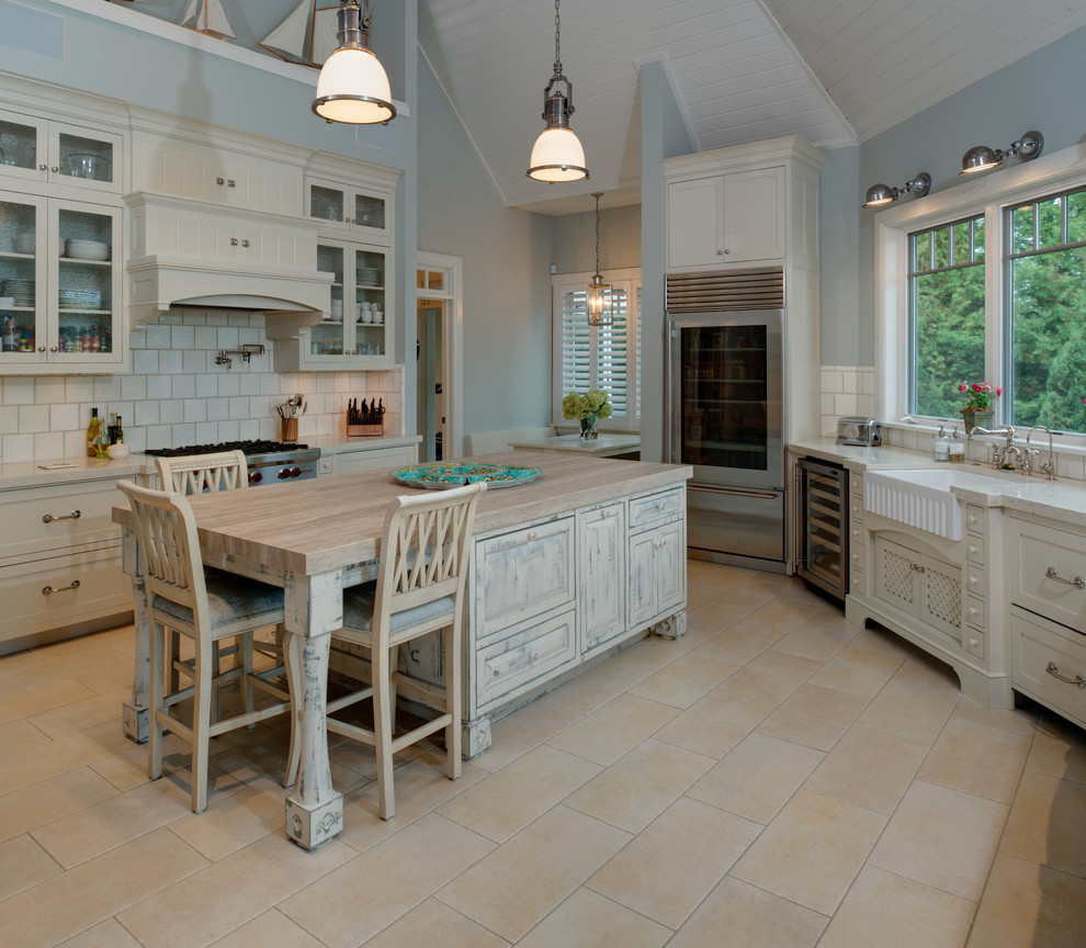 Photo of a large beach style u-shaped kitchen in Other with a farmhouse sink, recessed-panel cabinets, distressed cabinets, white splashback, stainless steel appliances, with island, beige floor, white benchtop, wood benchtops, porcelain floors and porcelain splashback.