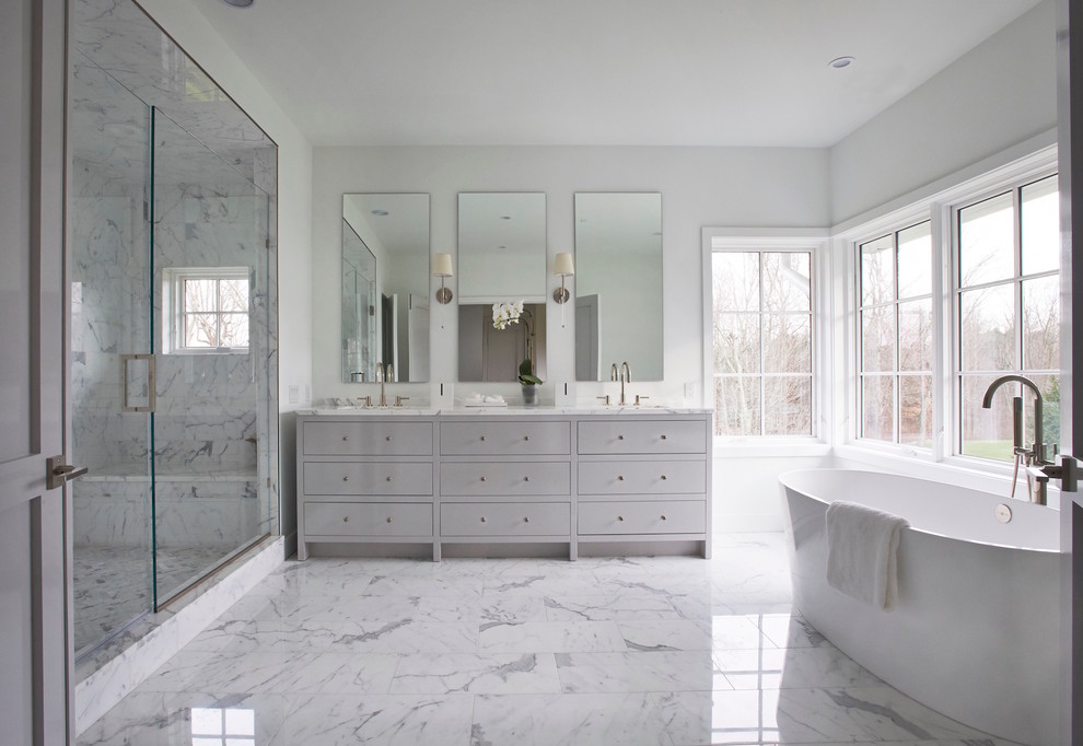 This is an example of a large transitional master bathroom in New York with grey cabinets, a freestanding tub, an alcove shower, white tile, stone tile, white walls, marble floors, an undermount sink, marble benchtops and flat-panel cabinets.