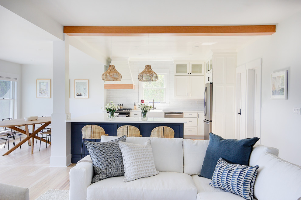 Photo of a mid-sized beach style open plan kitchen in Other with a farmhouse sink, recessed-panel cabinets, blue cabinets, quartz benchtops, white splashback, ceramic splashback, stainless steel appliances, light hardwood floors, with island, white benchtop and timber.