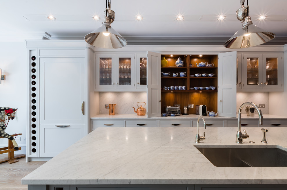 This is an example of a large transitional kitchen in London with grey cabinets, with island, marble benchtops, glass-front cabinets, stone slab splashback, light hardwood floors, beige floor and a double-bowl sink.