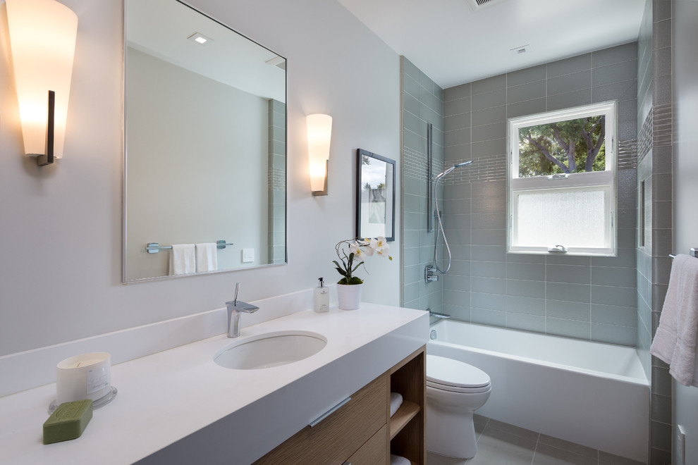 Design ideas for a mid-sized modern 3/4 bathroom in San Francisco with flat-panel cabinets, light wood cabinets, an alcove tub, an alcove shower, a one-piece toilet, green tile, white walls, engineered quartz benchtops, glass tile, a drop-in sink, porcelain floors and grey floor.