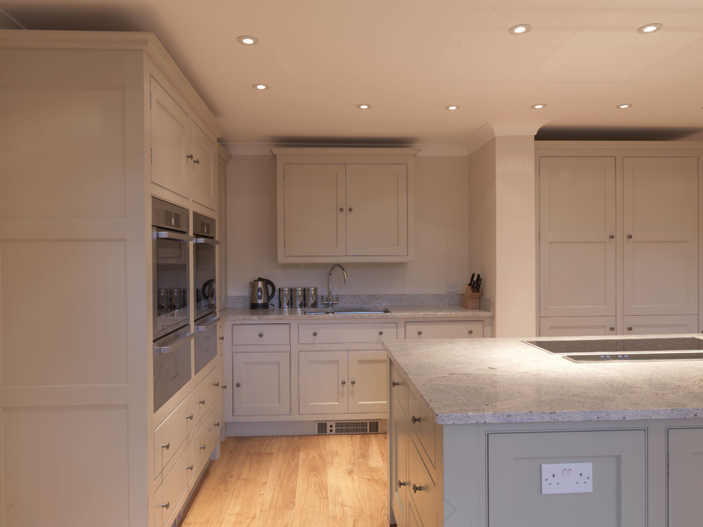 This is an example of a mid-sized traditional l-shaped open plan kitchen in Hampshire with a drop-in sink, shaker cabinets, beige cabinets, granite benchtops, stainless steel appliances, light hardwood floors and with island.