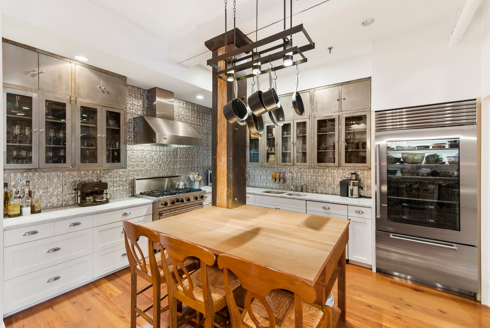 Photo of a transitional l-shaped kitchen in New York with a double-bowl sink, glass-front cabinets, stainless steel cabinets, metallic splashback, metal splashback, stainless steel appliances, medium hardwood floors and with island.