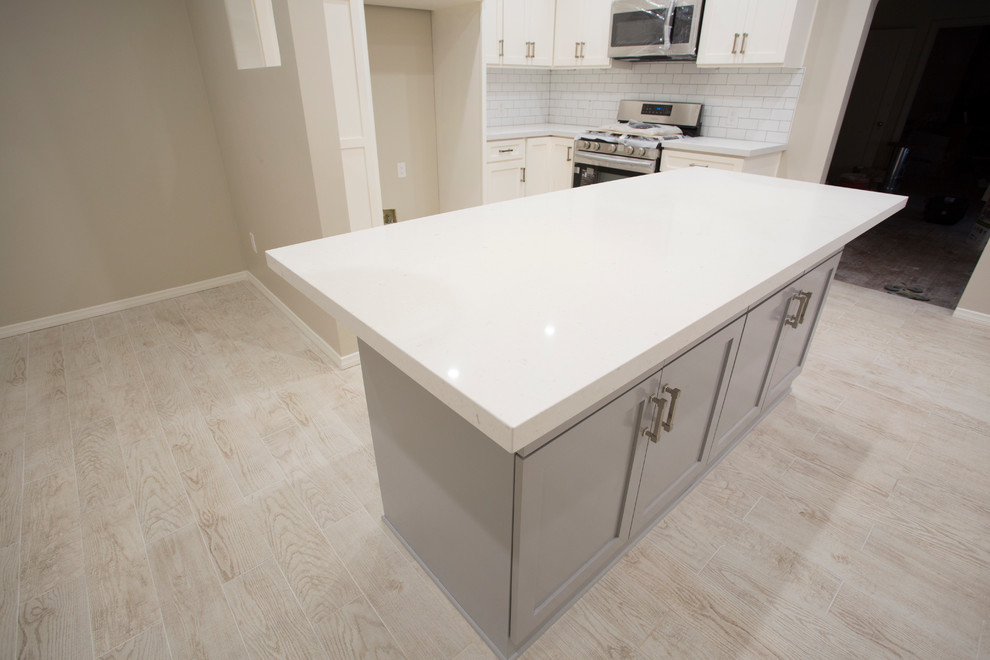 Photo of a mid-sized transitional single-wall open plan kitchen in Los Angeles with a farmhouse sink, shaker cabinets, white cabinets, quartzite benchtops, white splashback, subway tile splashback, stainless steel appliances, porcelain floors, with island, beige floor and white benchtop.