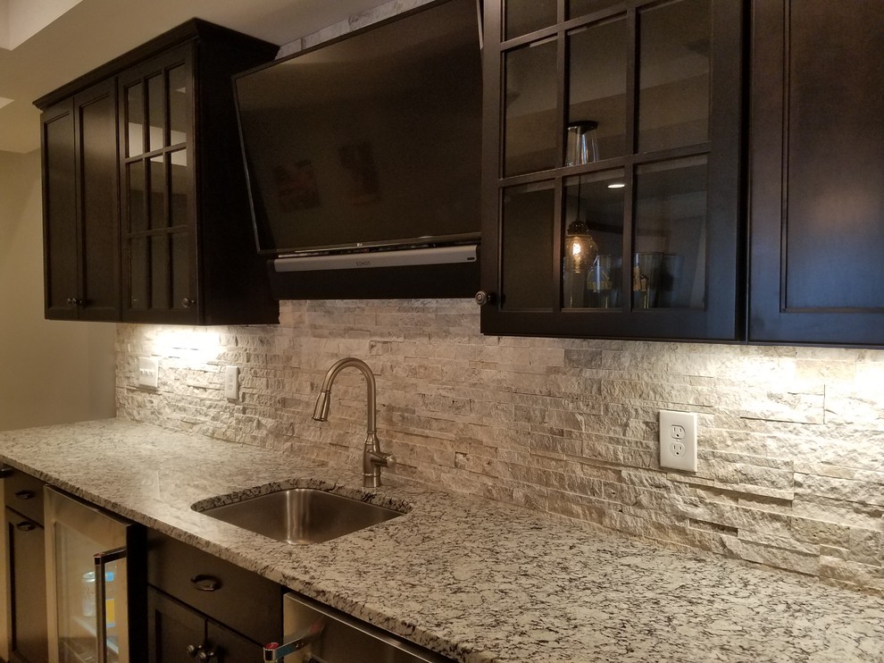 Inspiration for a mid-sized transitional single-wall wet bar in Cincinnati with a drop-in sink, flat-panel cabinets, dark wood cabinets, granite benchtops, grey splashback, stone tile splashback and brown floor.