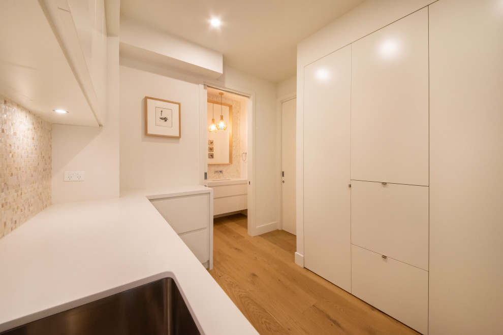 Inspiration for a large beach style l-shaped dedicated laundry room in Melbourne with an undermount sink, flat-panel cabinets, white cabinets, quartz benchtops, multi-coloured splashback, mosaic tile splashback, white walls, medium hardwood floors, a side-by-side washer and dryer and white benchtop.
