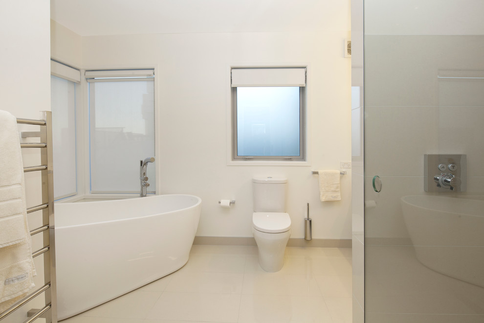 Photo of a large contemporary bathroom in Christchurch with a freestanding tub, an alcove shower, a one-piece toilet, white tile, porcelain tile, white walls, porcelain floors, white floor and a hinged shower door.
