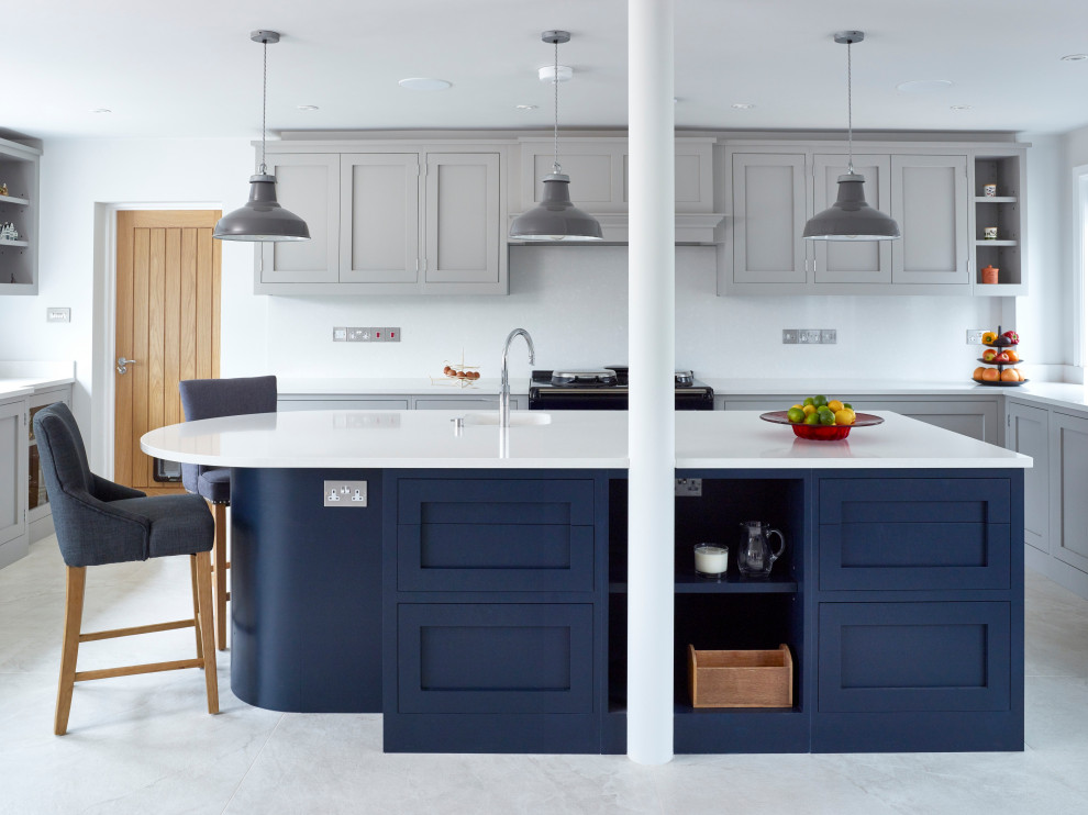 This is an example of a large transitional open plan kitchen in Oxfordshire with an integrated sink, shaker cabinets, blue cabinets, quartzite benchtops, white splashback, stone slab splashback, black appliances, porcelain floors, with island, white floor and white benchtop.
