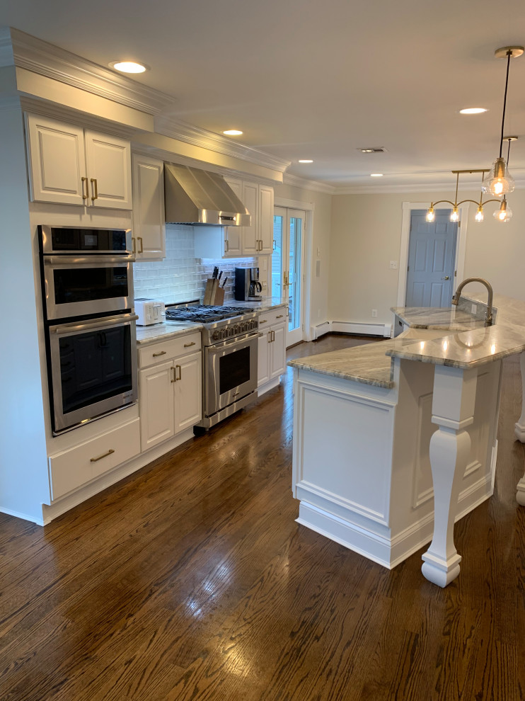 Large transitional l-shaped medium tone wood floor and brown floor open concept kitchen photo with a farmhouse sink, raised-panel cabinets, white cabinets, granite countertops, white backsplash, ceramic backsplash, stainless steel appliances, an island and gray countertops