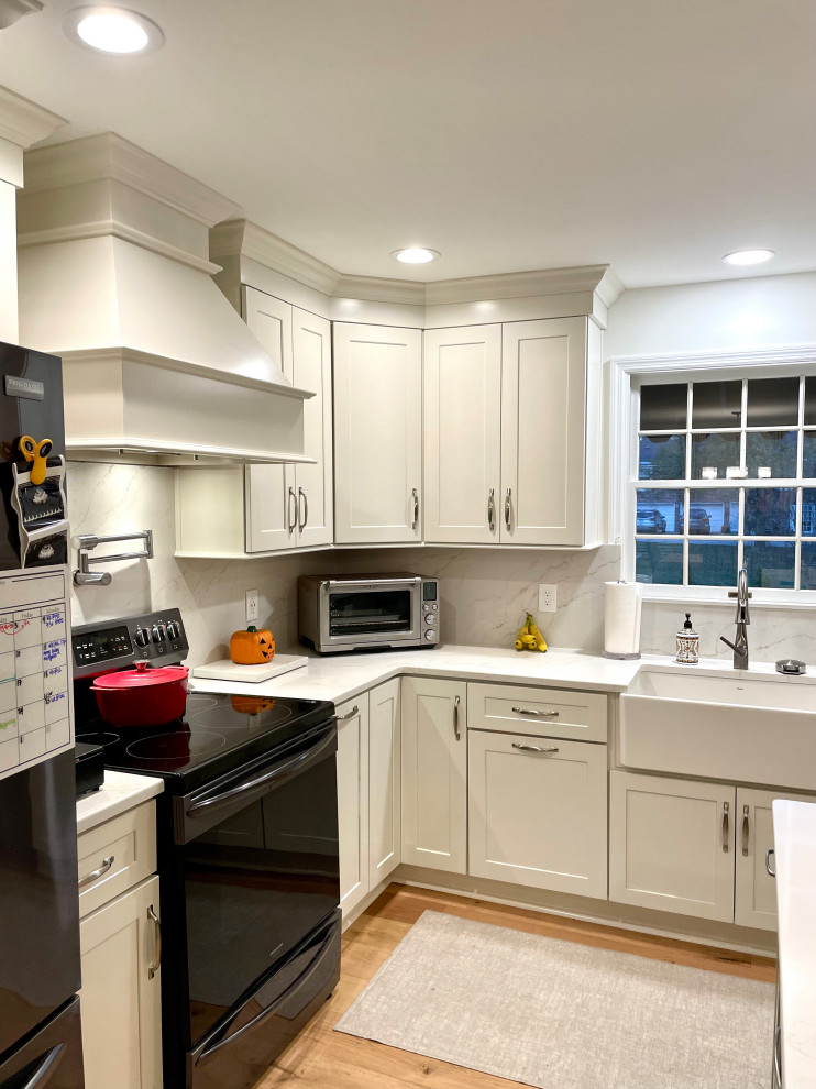 Transitional l-shaped light wood floor and beige floor eat-in kitchen photo with a farmhouse sink, shaker cabinets, white cabinets, quartz countertops, white backsplash, quartz backsplash, black appliances, an island and white countertops