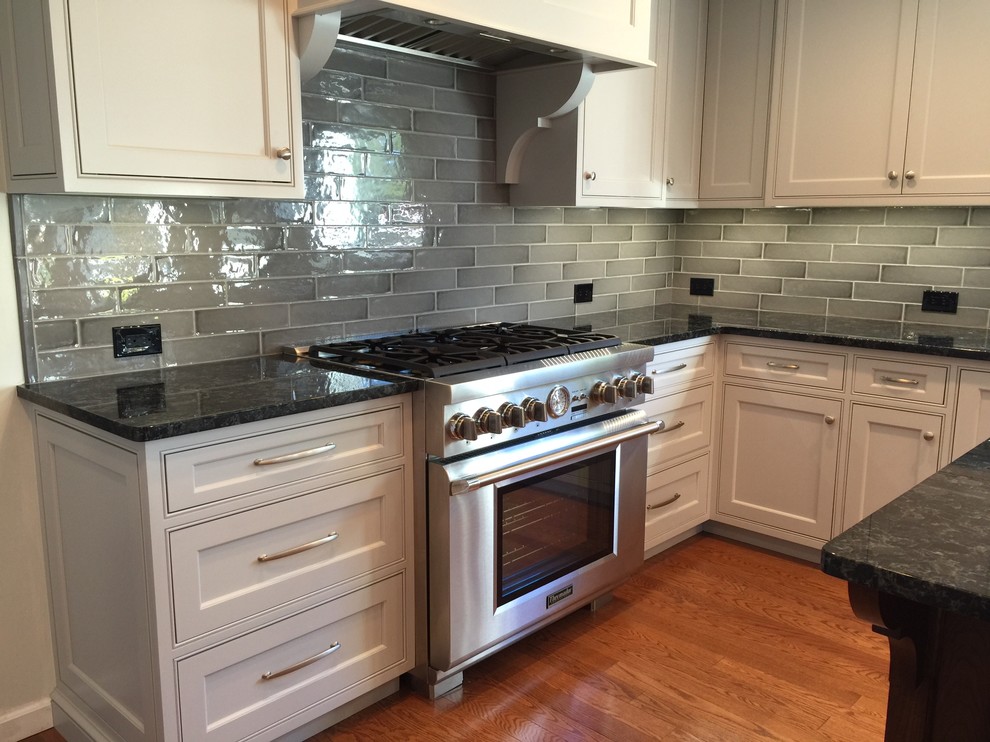 Photo of a large contemporary l-shaped eat-in kitchen in New York with a farmhouse sink, flat-panel cabinets, grey cabinets, granite benchtops, grey splashback, ceramic splashback, stainless steel appliances, medium hardwood floors and with island.