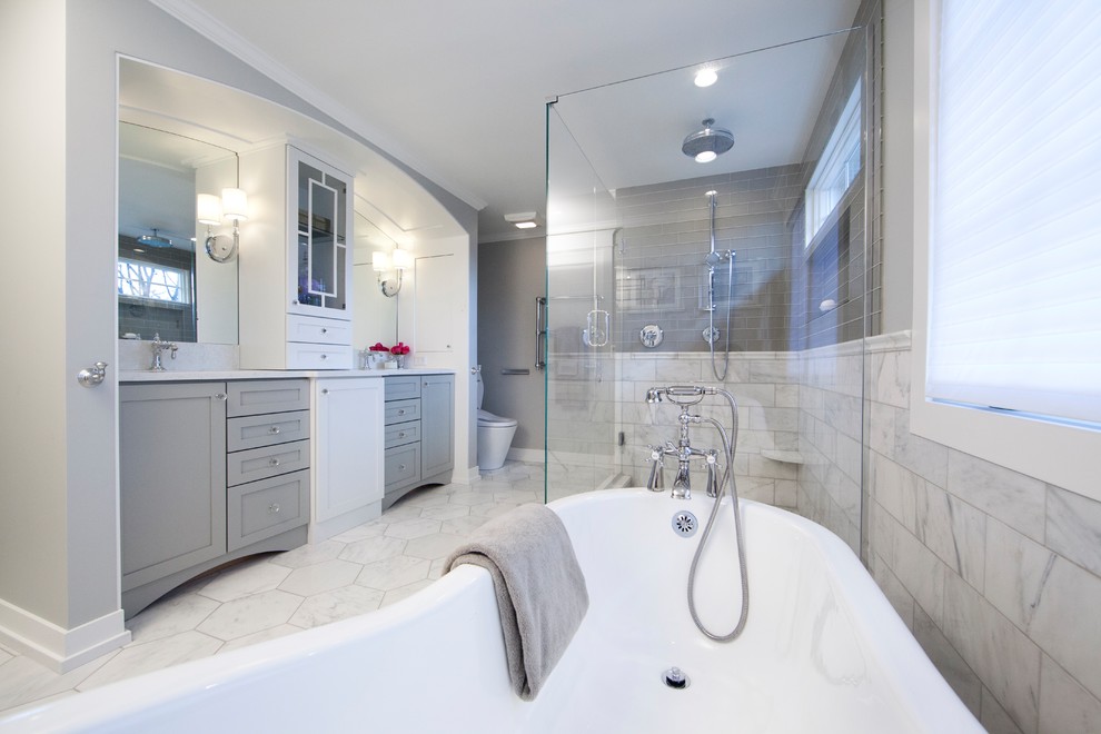This is an example of a large traditional master bathroom in Grand Rapids with shaker cabinets, grey cabinets, a freestanding tub, a corner shower, a one-piece toilet, white tile, marble, grey walls, marble floors, an undermount sink, marble benchtops, white floor and a hinged shower door.