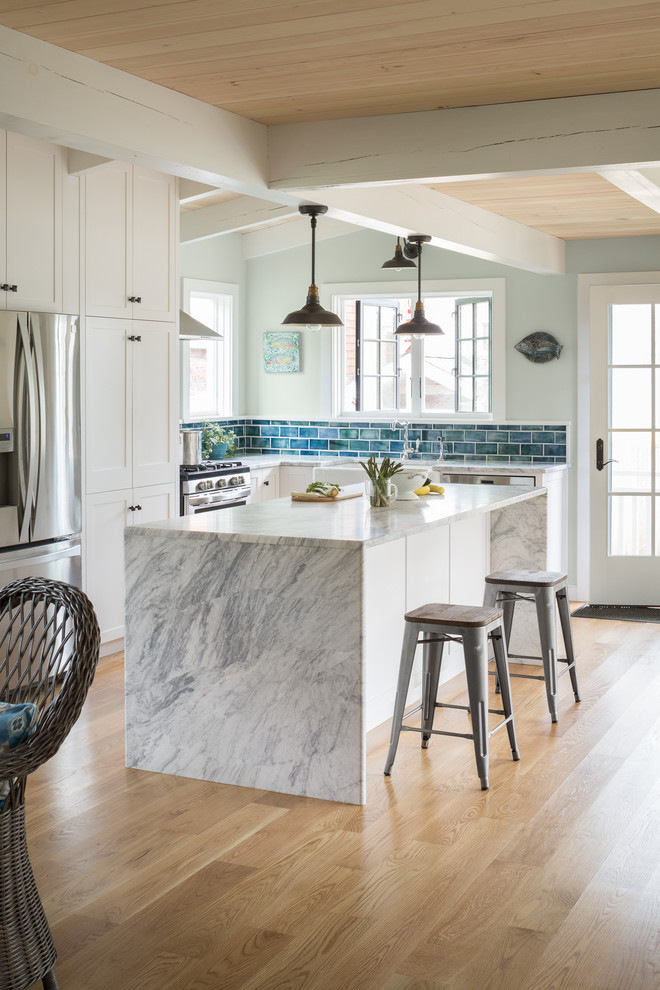 This is an example of a beach style l-shaped open plan kitchen in Portland Maine with shaker cabinets, white cabinets, blue splashback, stainless steel appliances, medium hardwood floors, with island and brown floor.