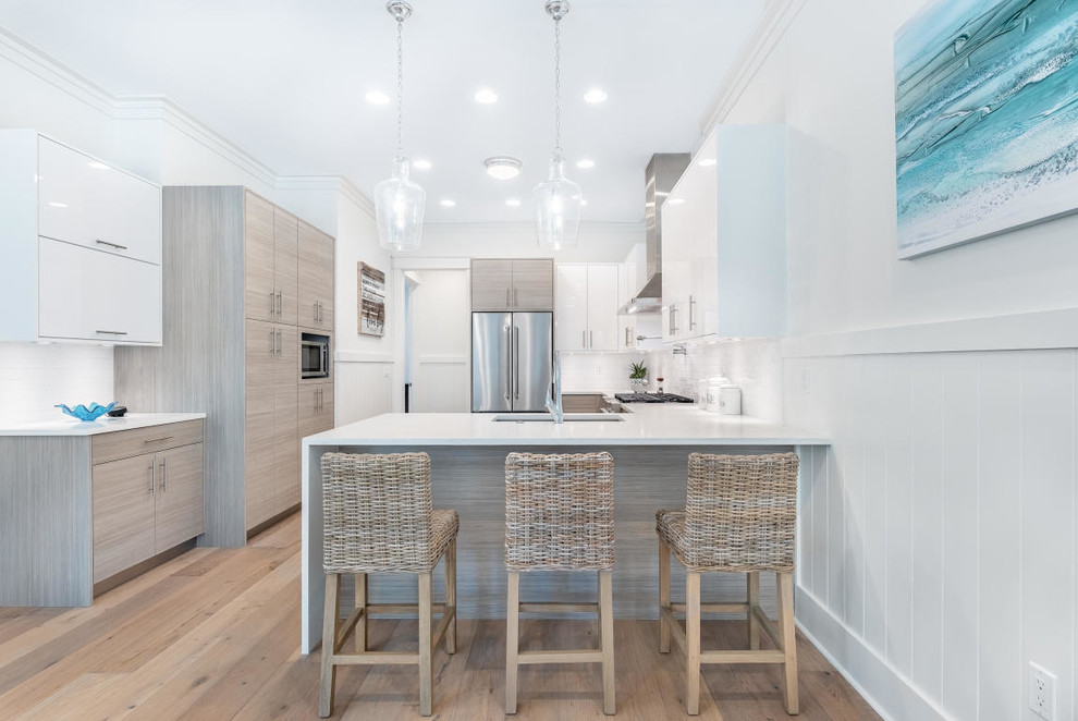 Photo of a mid-sized beach style u-shaped eat-in kitchen in Miami with an undermount sink, flat-panel cabinets, white cabinets, quartz benchtops, white splashback, subway tile splashback, stainless steel appliances, light hardwood floors, a peninsula, brown floor and white benchtop.