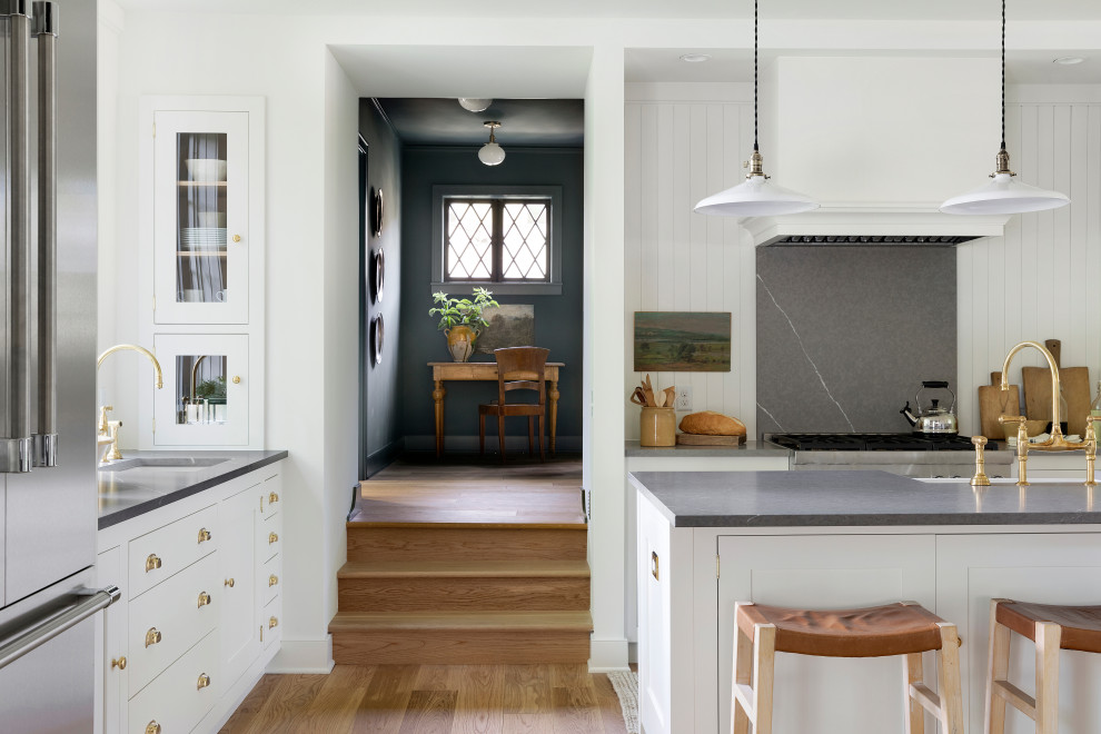 Foto på ett mellanstort lantligt grå kök, med en rustik diskho, skåp i shakerstil, vita skåp, bänkskiva i kvarts, vitt stänkskydd, rostfria vitvaror, ljust trägolv, en köksö och brunt golv