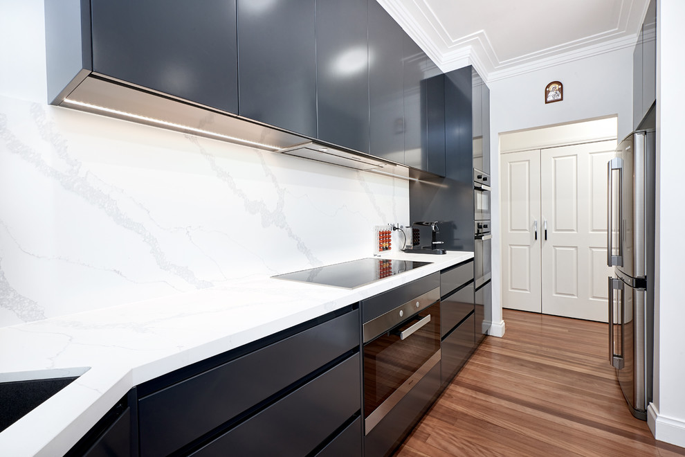 Mid-sized modern u-shaped kitchen pantry in Melbourne with an undermount sink, black cabinets, solid surface benchtops, white splashback, stone slab splashback, stainless steel appliances, light hardwood floors, brown floor and white benchtop.