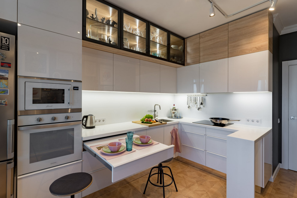 This is an example of a contemporary l-shaped kitchen in Saint Petersburg with an undermount sink, flat-panel cabinets, white cabinets, white appliances, medium hardwood floors, a peninsula, brown floor and white benchtop.