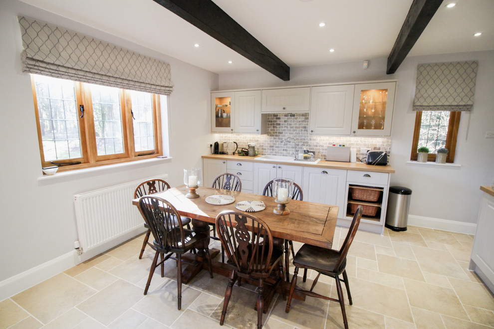 Mid-sized country l-shaped eat-in kitchen in London with an integrated sink, shaker cabinets, grey cabinets, wood benchtops, multi-coloured splashback, brick splashback, black appliances, limestone floors and beige floor.