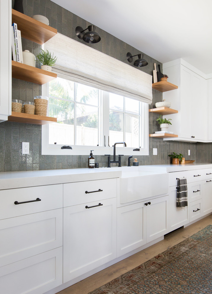 Example of a mid-sized transitional l-shaped light wood floor and beige floor kitchen design in San Diego with a farmhouse sink, shaker cabinets, white cabinets, solid surface countertops, gray backsplash, subway tile backsplash, paneled appliances and an island