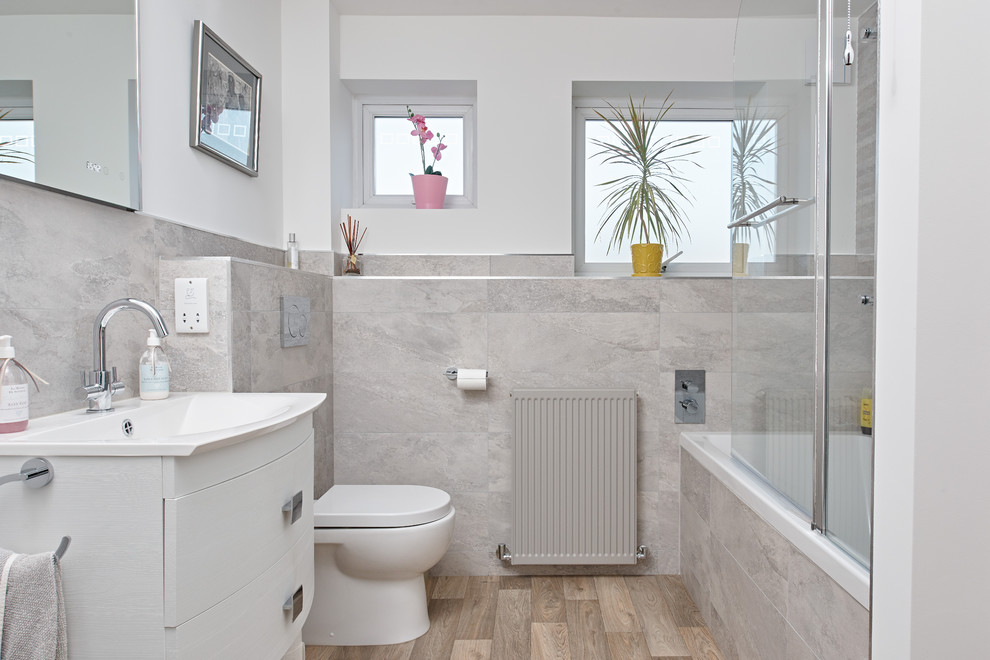 Photo of a mid-sized transitional kids bathroom in Berkshire with flat-panel cabinets, white cabinets, a drop-in tub, a shower/bathtub combo, a one-piece toilet, gray tile, ceramic tile, white walls, vinyl floors, a wall-mount sink, brown floor and a hinged shower door.