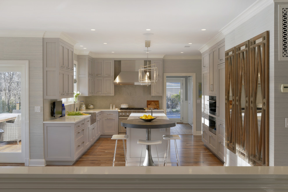 Photo of an expansive traditional u-shaped eat-in kitchen in New York with a farmhouse sink, shaker cabinets, grey cabinets, glass benchtops, grey splashback, limestone splashback, panelled appliances, medium hardwood floors, with island and brown floor.